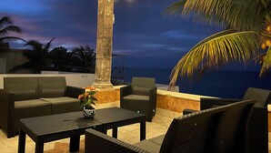 Un espace extérieur aménagé avec des meubles en osier et une colonne centrale, offrant une vue sur une plage au crépuscule.