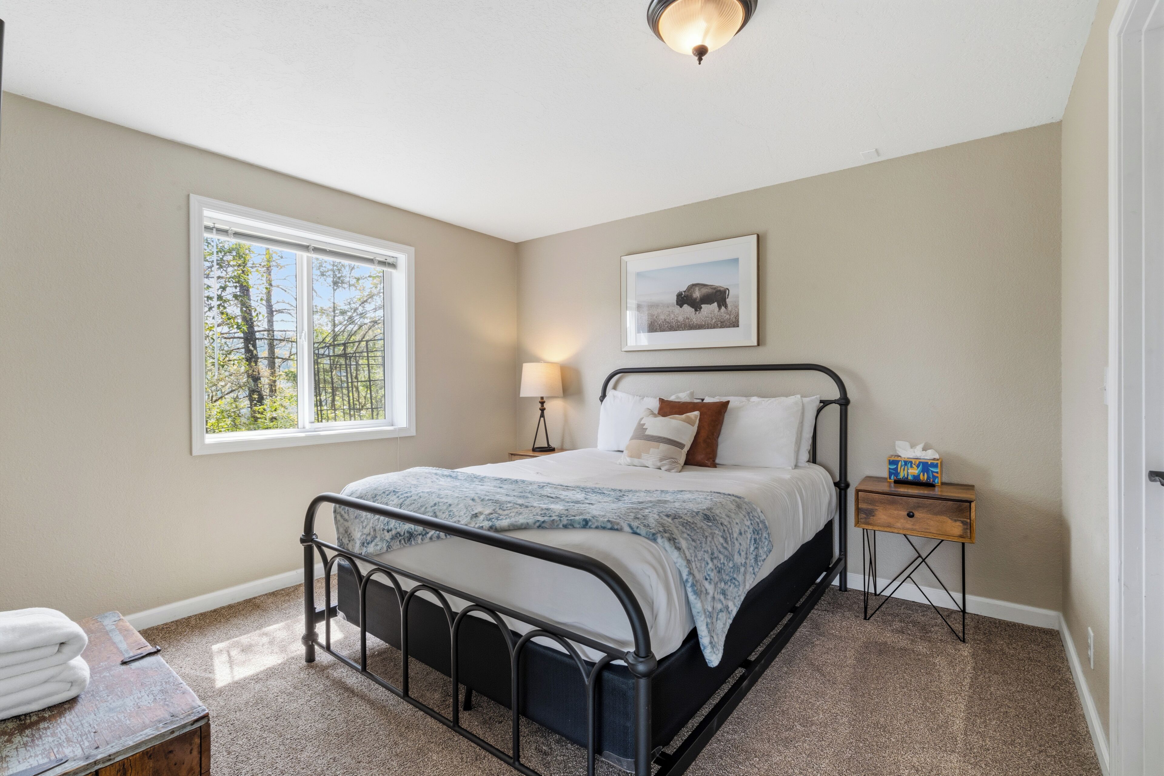 A bedroom with a bed, a bedside table, a lamp, a framed picture on the wall, and a window with a view of trees.