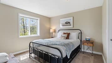 A bedroom with a bed, a bedside table, a lamp, a framed picture on the wall, and a window with a view of trees.