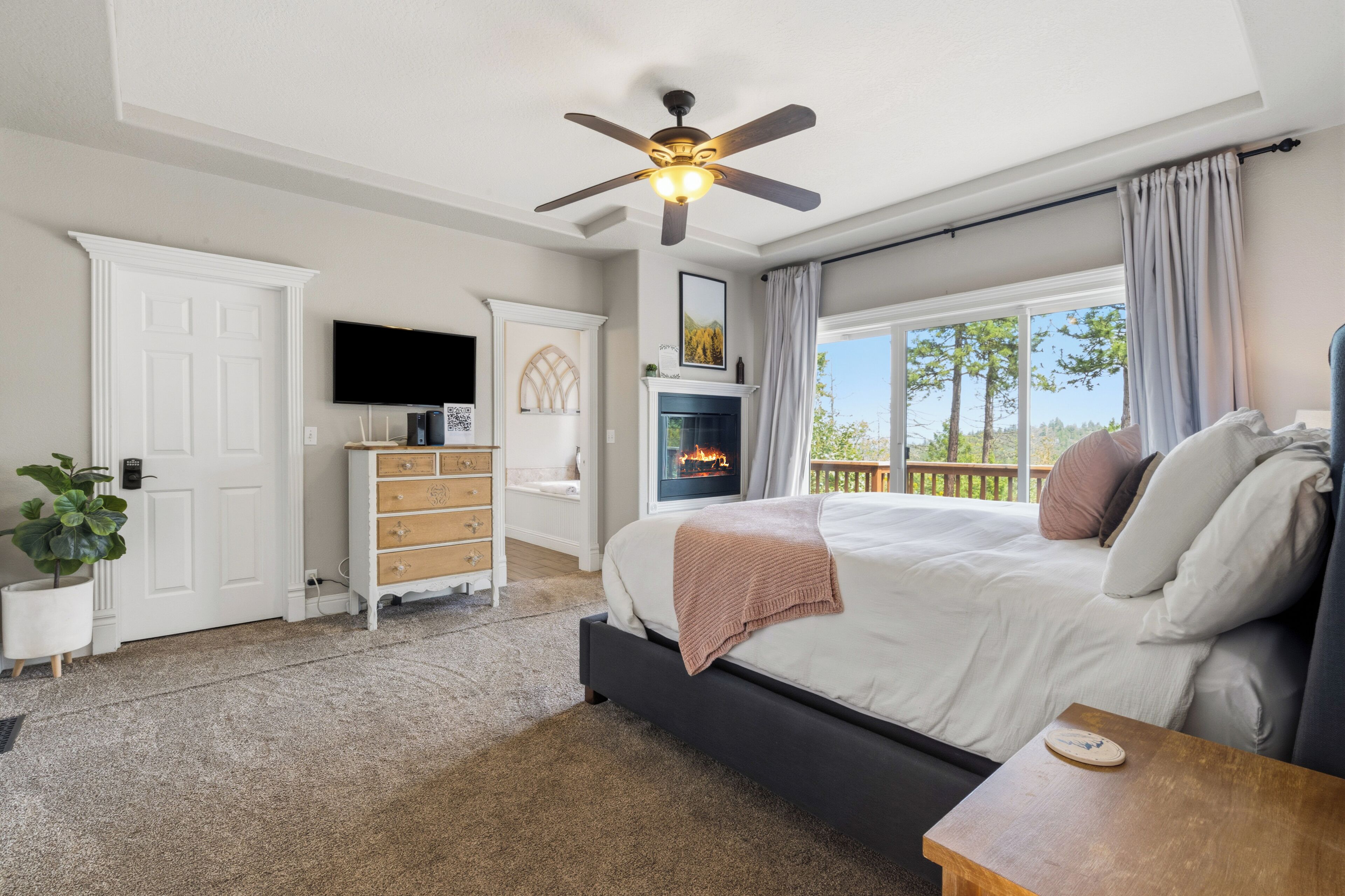 A bedroom with a bed, a ceiling fan, a fireplace, a TV, and a view of the outdoors.