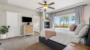A bedroom with a bed, a ceiling fan, a fireplace, a TV, and a view of the outdoors.