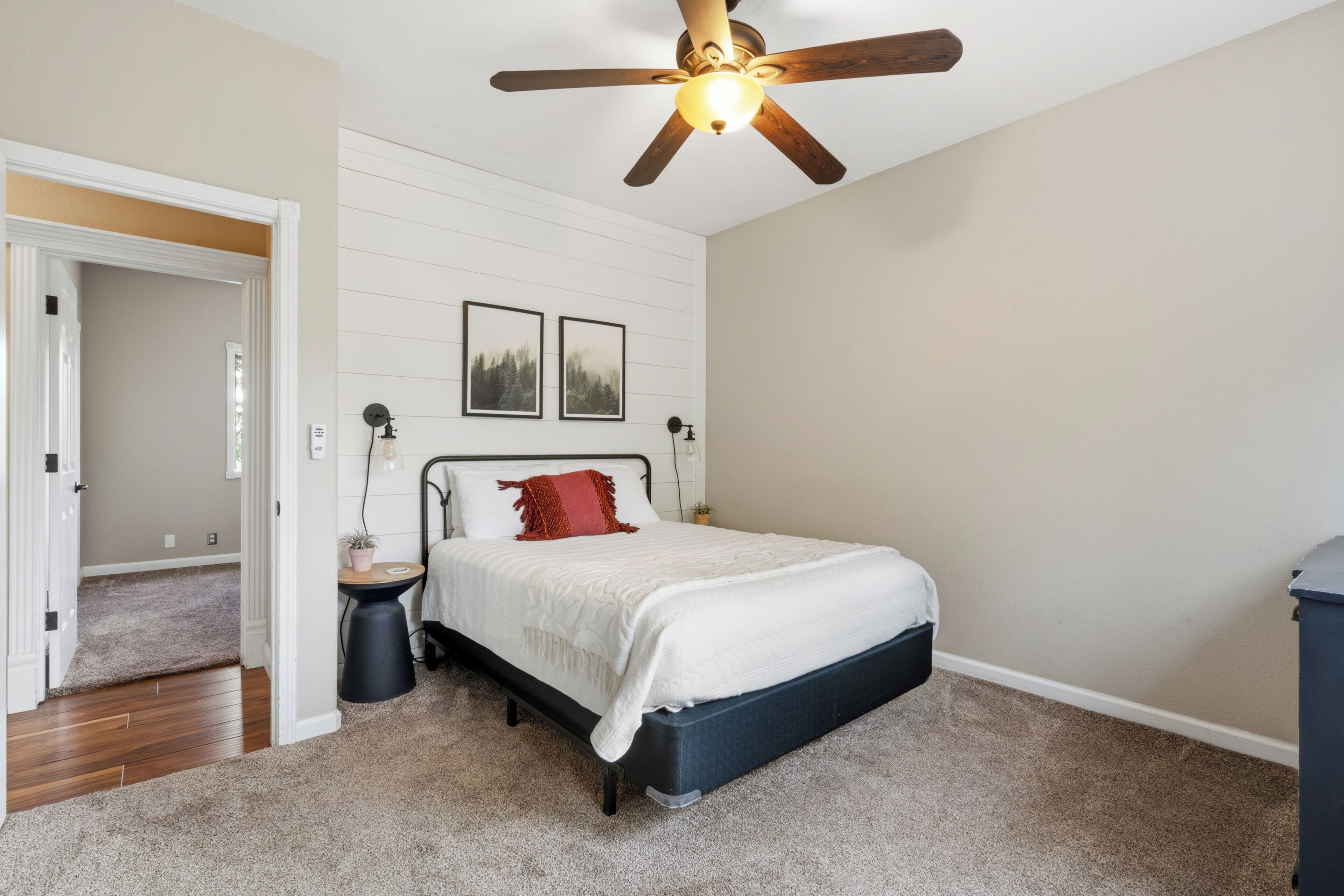 A bedroom with a bed, a ceiling fan, a nightstand, a wall-mounted lamp, and two framed pictures.