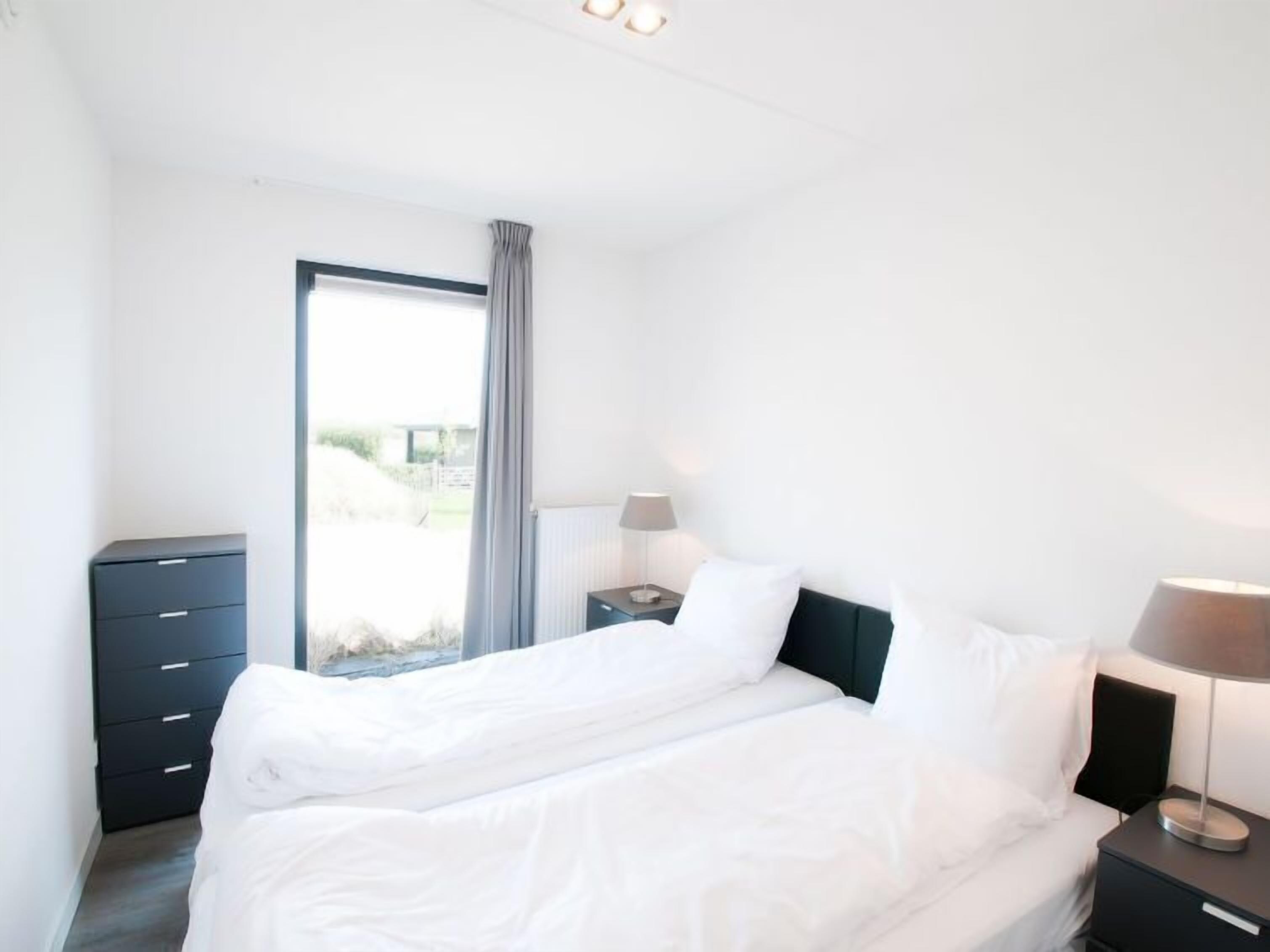 Une chambre à coucher moderne avec un grand lit, une table de chevet, une commode et une fenêtre avec des rideaux.