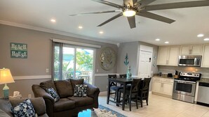 A living room with a brown sofa, a coffee table, and a ceiling fan. There is a dining area with a table and chairs, and a kitchen with cabinets and appliances.