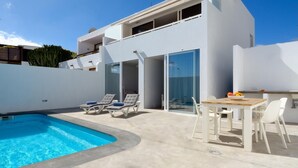 Una villa moderna con piscina, lettini prendisole, zona pranzo e una porta a vetri che dà su un balcone.