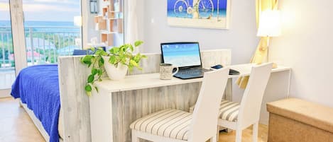A bedroom with a bed, a desk with a laptop, a chair, a plant, and a painting.