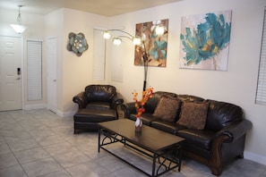 A living room with a dark leather sofa, a matching armchair, a coffee table, and a vase with flowers. There are two wall-mounted light fixtures and a large abstract painting.