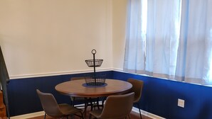 Un comedor con una mesa redonda, cuatro sillas, un soporte de metal negro y una ventana con cortinas translúcidas.