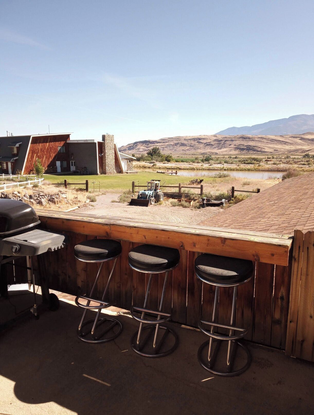 Un coin bar avec trois tabourets, un barbecue et une vue sur un paysage rural avec des montagnes et une étendue d’eau.