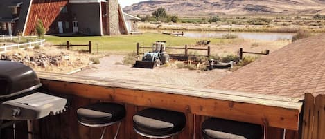 Un espace bar avec des tabourets et un grill, offrant une vue panoramique sur un paysage magnifique, avec des montagnes et une étendue d’eau.