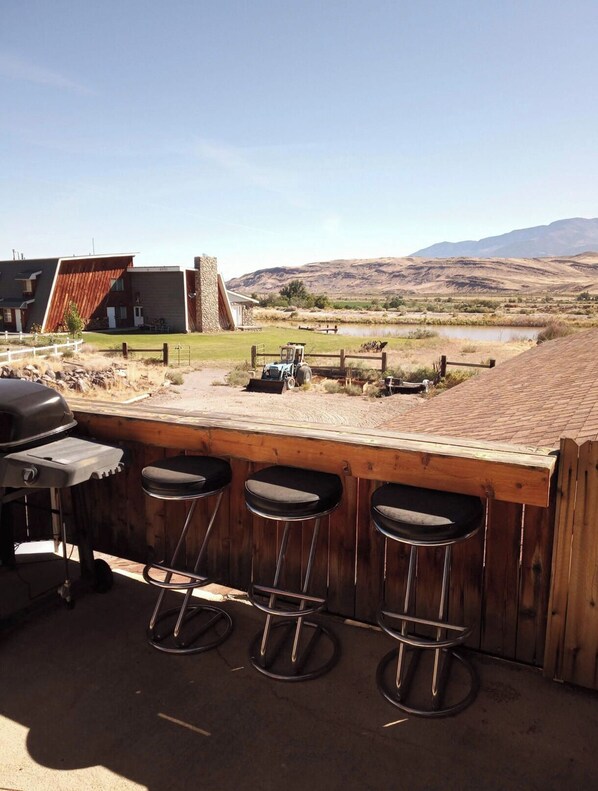 Zona de bar con tres taburetes, una parrilla y vistas a un paisaje rural con montañas y una masa de agua.