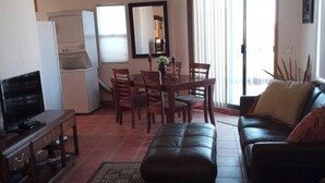 Een woonkamer met een televisie, een bank, een eettafel met stoelen, een plafondventilator en een raam met gordijnen.