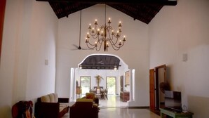 A spacious living room with a high ceiling, a chandelier, a sofa, a television, and a dining area in the background.