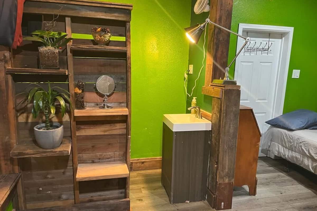 A room with a green wall, wooden shelving, a small sink, a mirror, a fan, and a bed with a blue pillow.