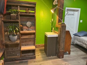 A room with a green wall, wooden shelving, a small sink, a mirror, a fan, and a bed with a blue pillow. - Dream catcher (Emeryville)