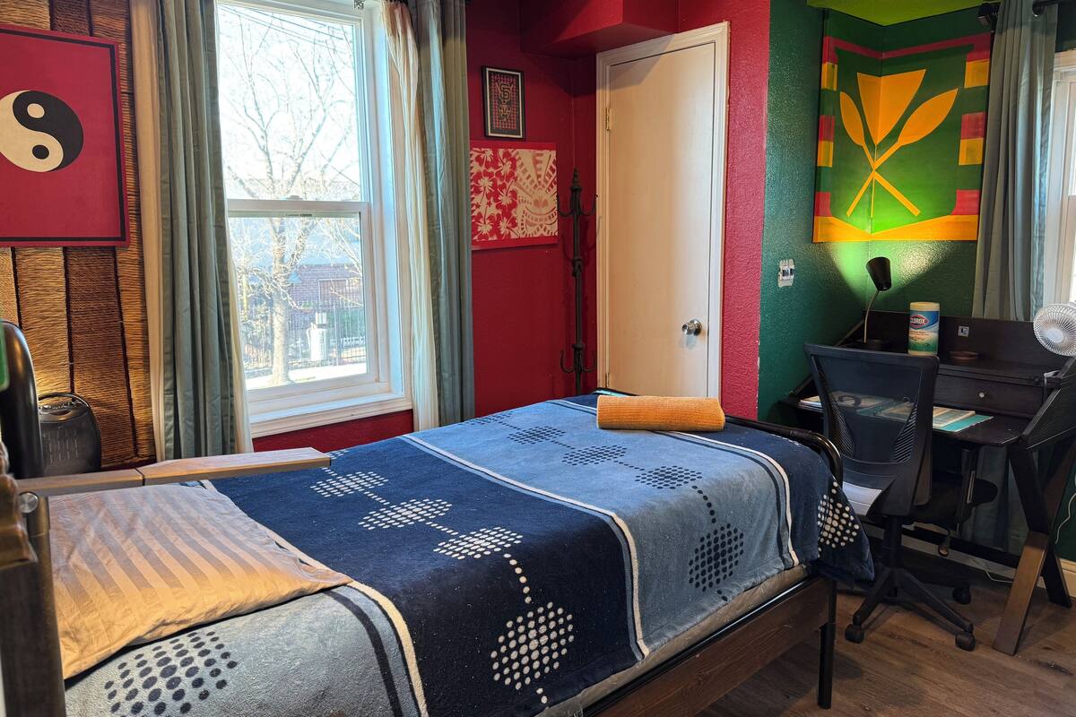 A room with a bed, a desk, and a chair. There is a window with curtains, a door, and a wall with a colorful mural.