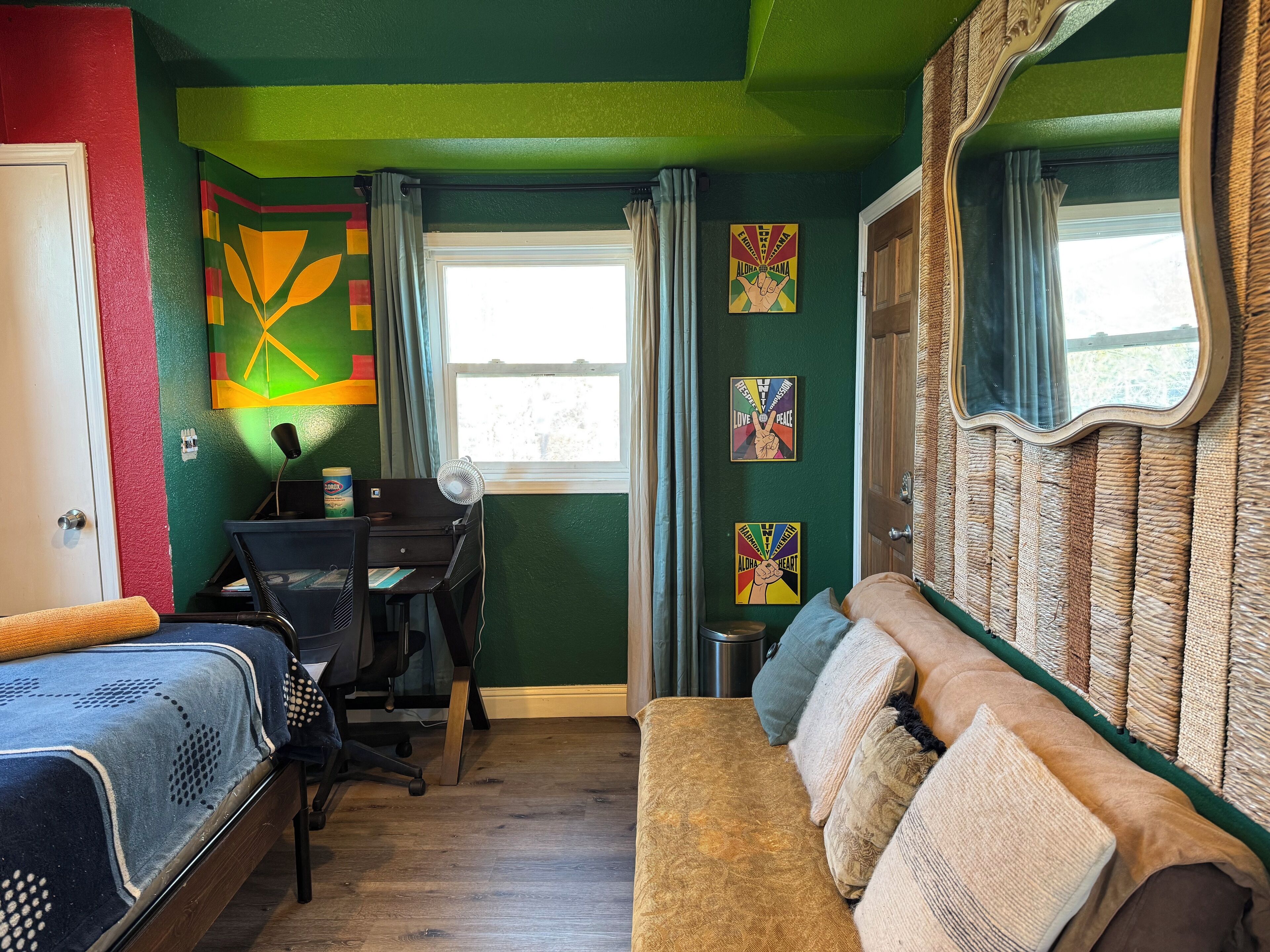Quarto com cama, escrivaninha e sofá. Possui duas janelas com cortinas, e as paredes são pintadas de verde.