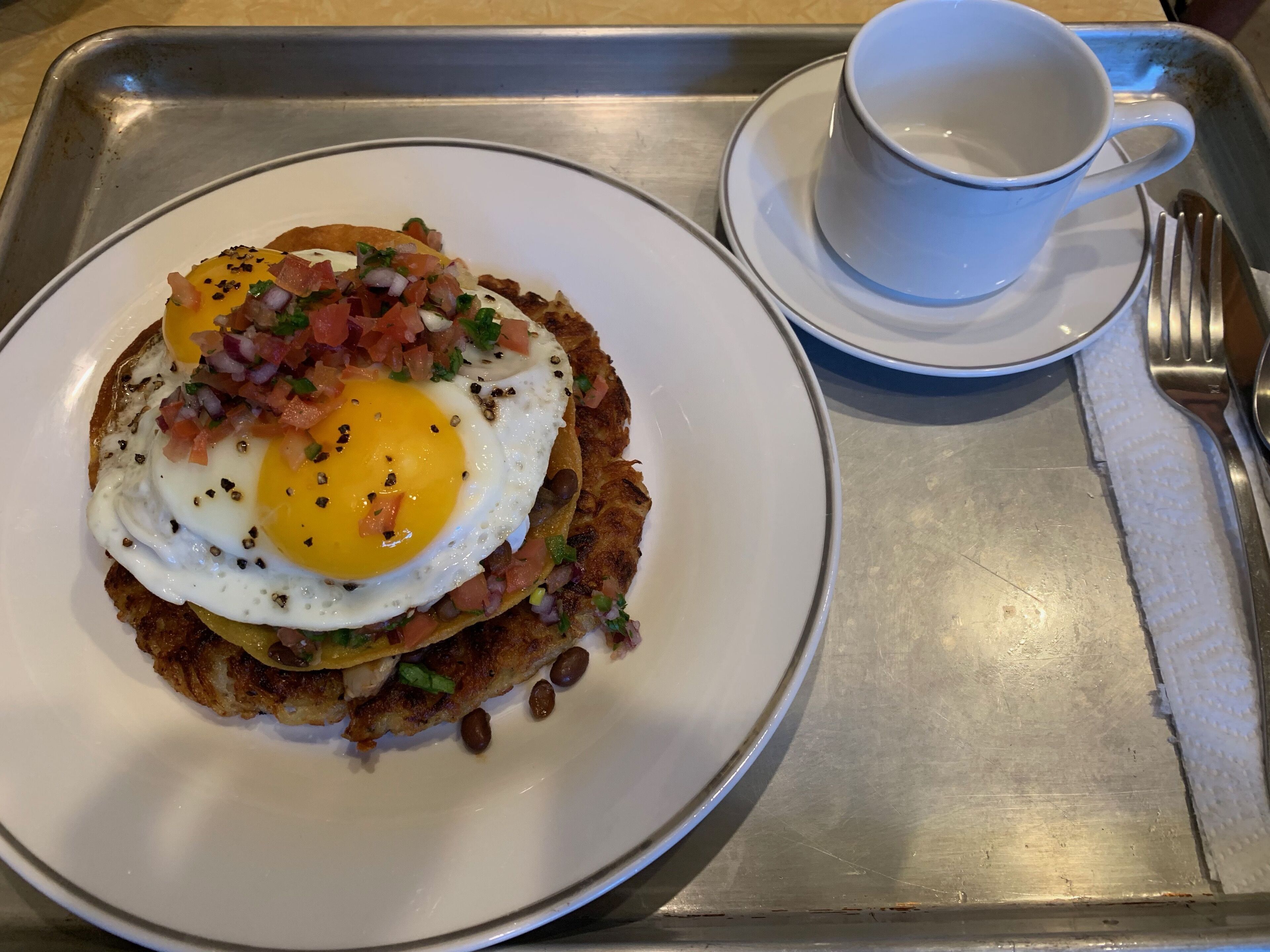 Ein Frühstücksbrett mit einer Portion Rösti, belegt mit einem Spiegelei und gewürfelten Tomaten, einer Kaffeetasse und einer Gabel.