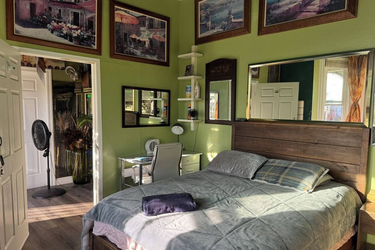 A bedroom with a bed, a desk, a chair, a mirror, and artwork on the walls.