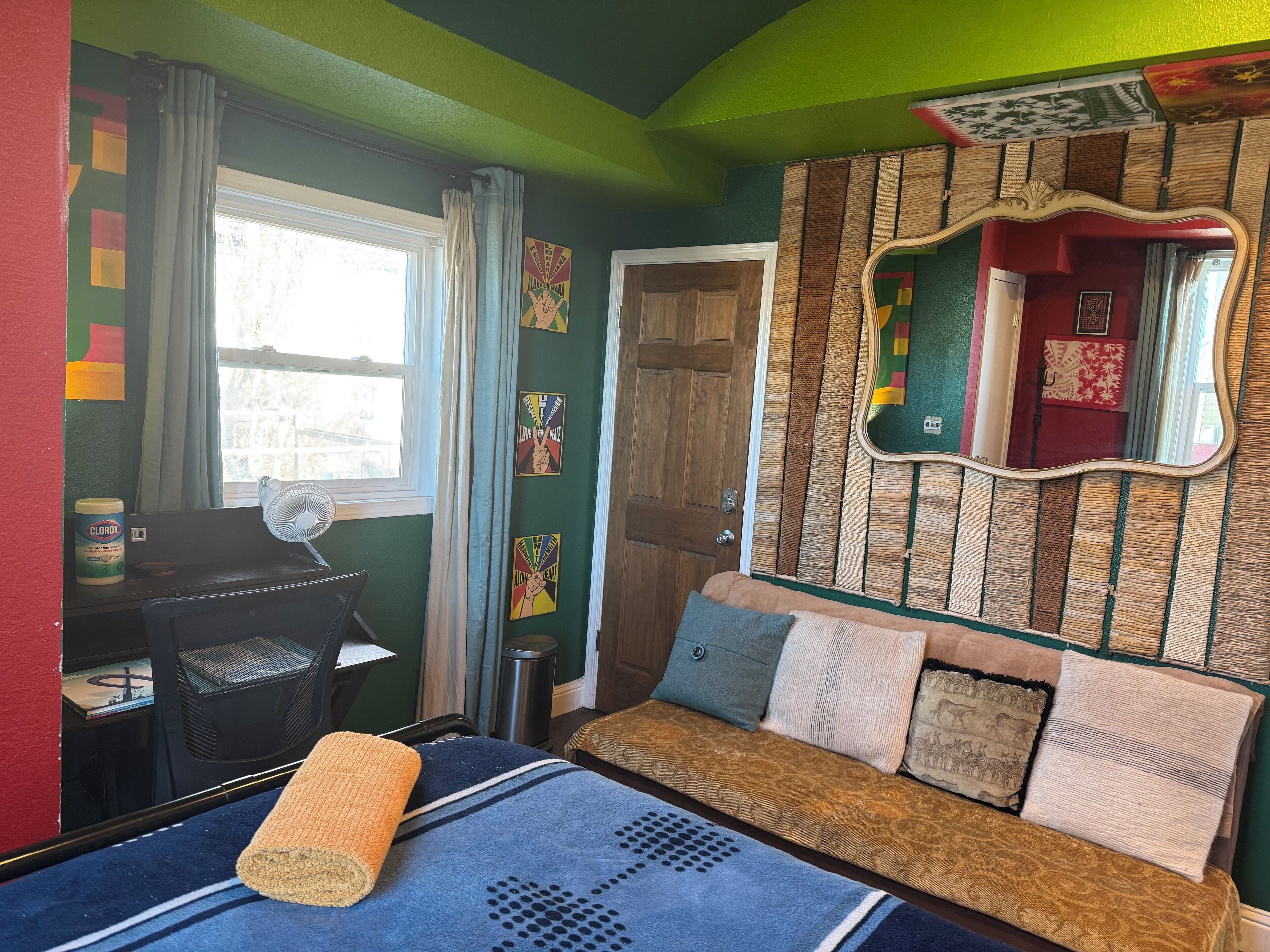 A room with a sofa, a fan, a window with curtains, a door, and a mirror with a wooden frame.