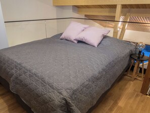 Ein Bett mit grauer gesteppter Bettdecke und zwei Kissen, ein Holz-Kopfschild und ein kleiner Tisch mit Lampe.