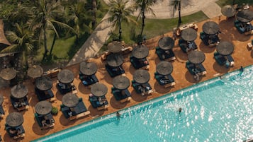 An aerial view of a pool area with lounge chairs and umbrellas, surrounded by palm trees and a sandy area.