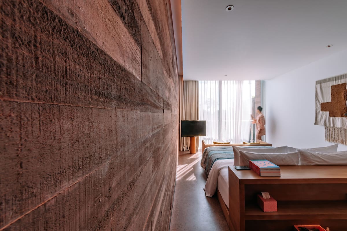 A modern hotel room with a large wooden accent wall, a bed with a white comforter, a bedside table with books and a tissue box, a flat-screen TV, and a window with sheer curtains.