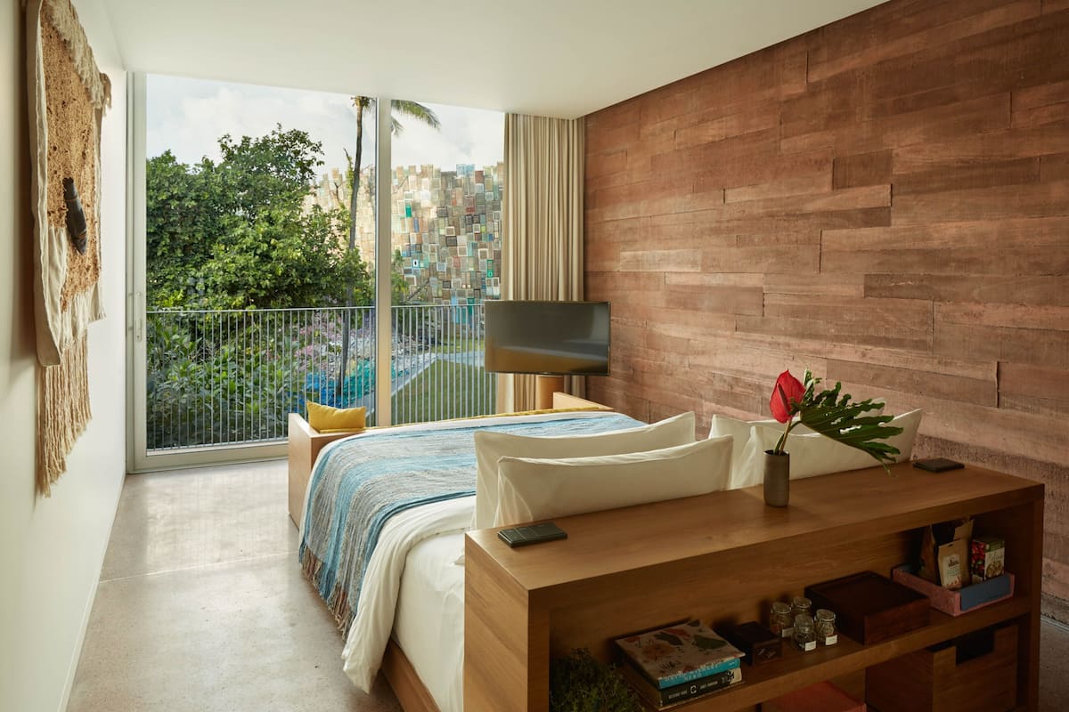 A modern bedroom with a large bed, a wooden bedside table, a TV, and a balcony with a view of buildings and greenery.