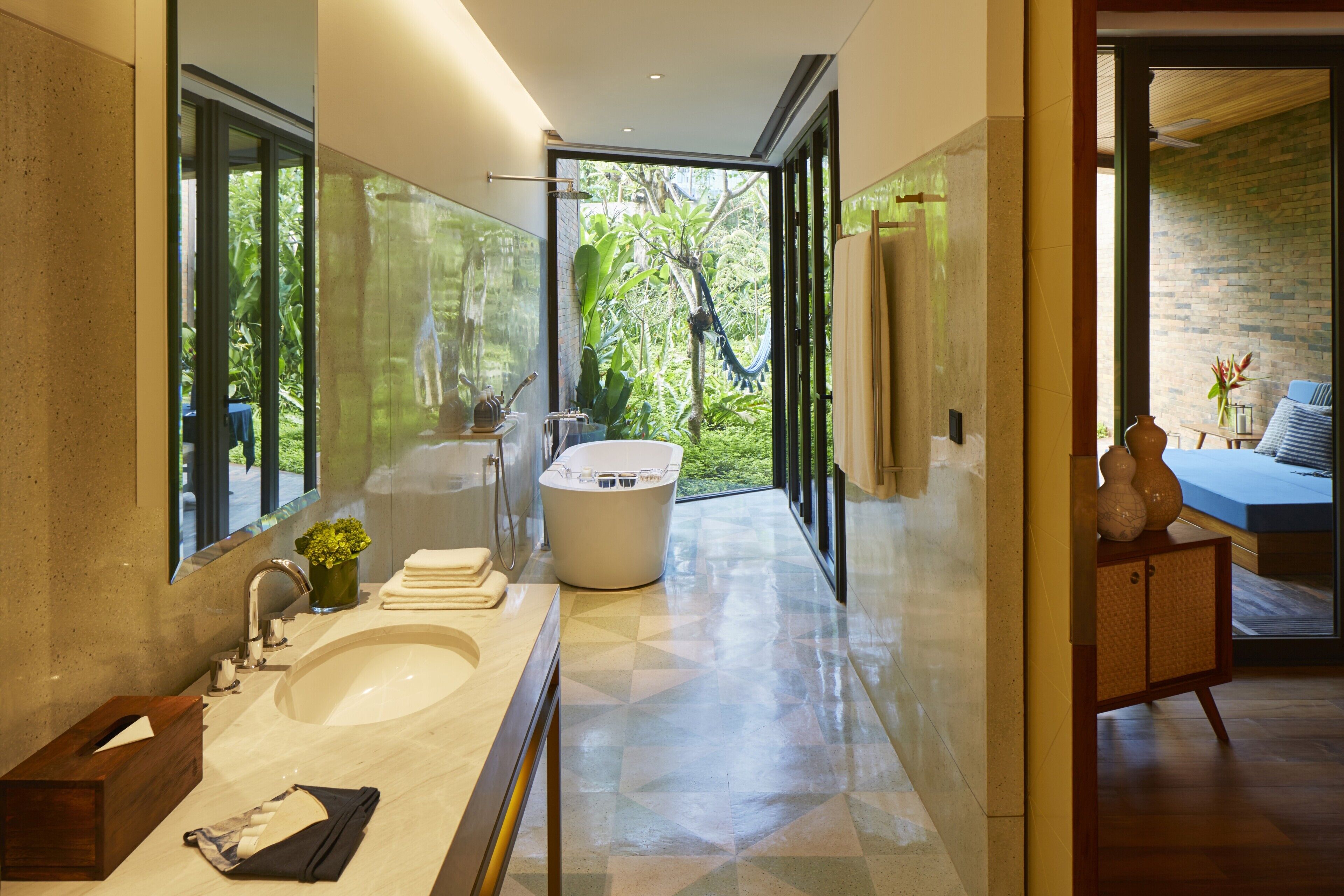 A modern bathroom with a large bathtub, a sink, and a mirror. There is a window providing a view of greenery outside.