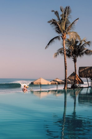 A poolside view with palm trees, a beach, and a person relaxing.