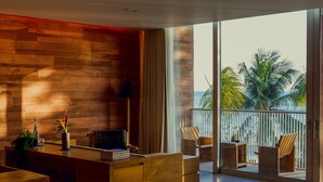 A modern living room with a wooden wall, a glass door leading to a balcony with a view of palm trees and the ocean. - Desa Potato Head Bali (Keroboken Kelod)