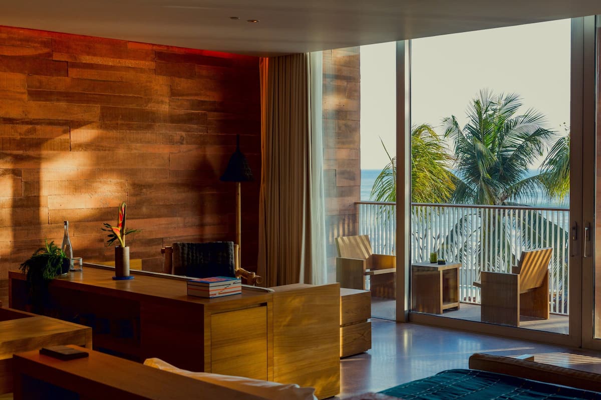 A modern living room with a wooden wall, a glass door leading to a balcony with a view of palm trees and the ocean.