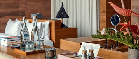 Una habitación de hotel con una cama de madera, una mesita de noche de madera con un neceser, un pequeño espejo y una planta en maceta.