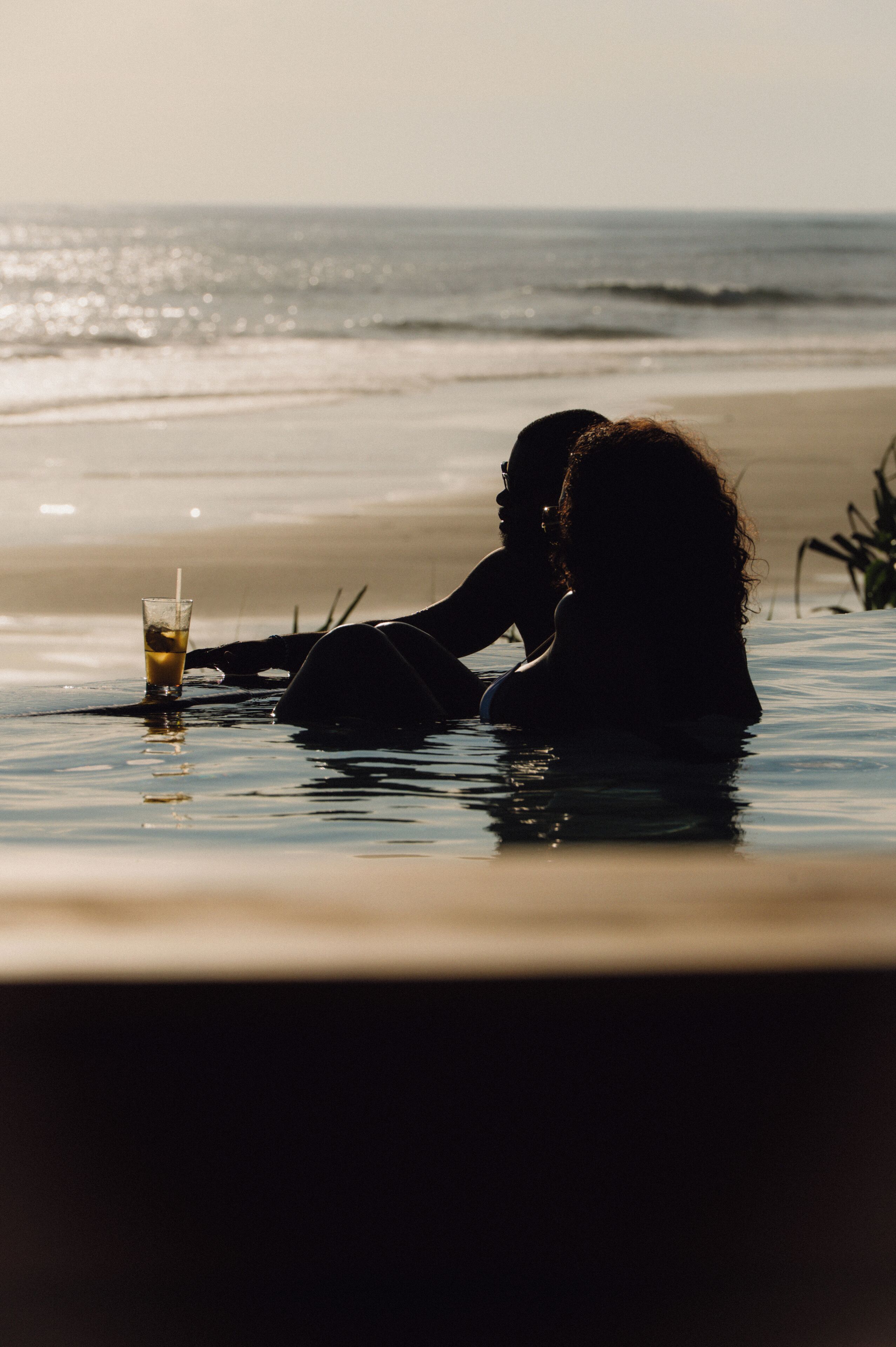 Um casal sentado à beira da piscina com uma bebida na mesa.