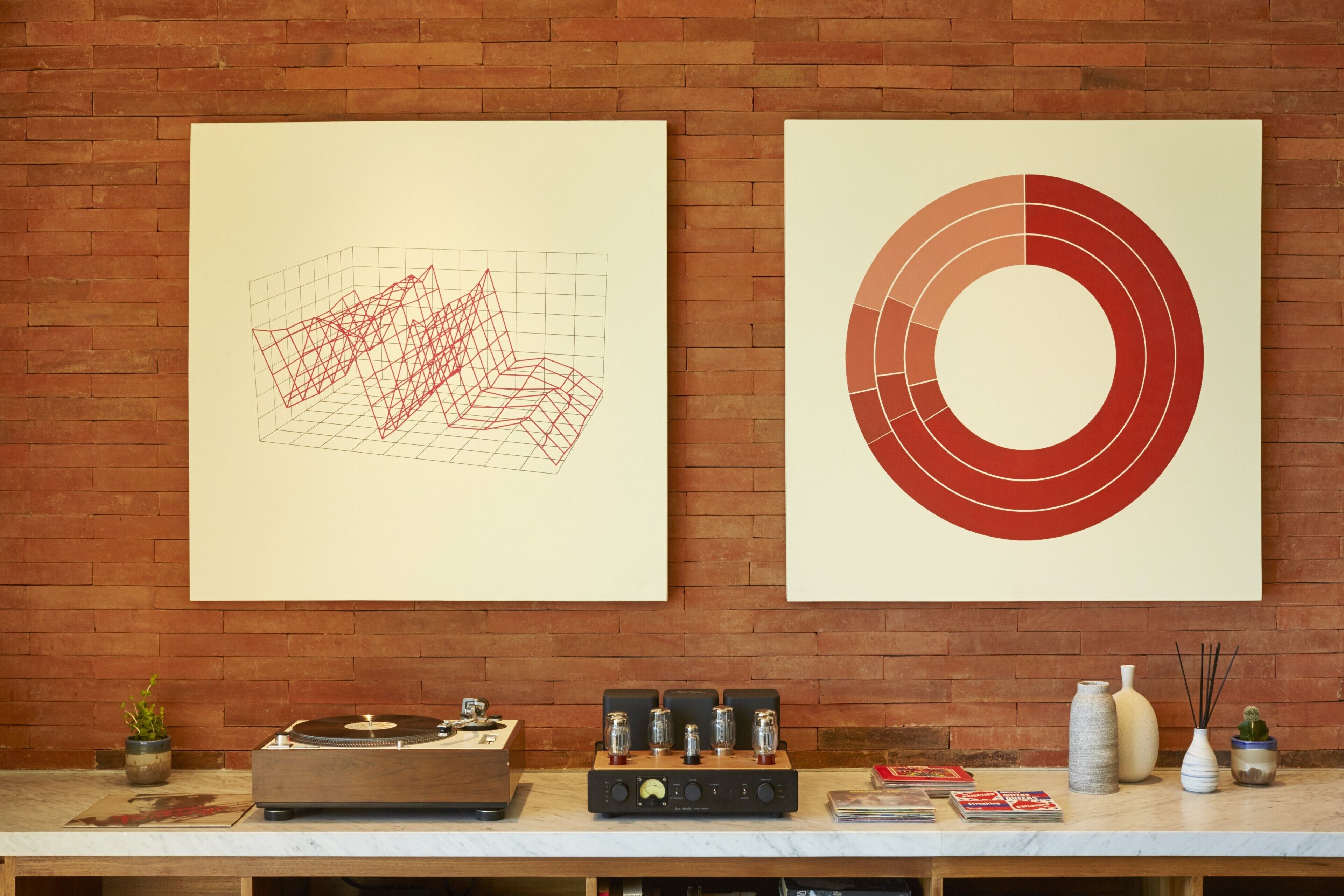 Two abstract paintings on a brick wall, a turntable with speakers, and a table with books and decorative items.