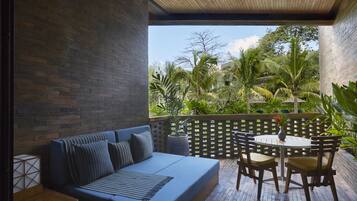 A modern outdoor seating area with a blue sofa and wooden chairs, a small table, and a view of lush greenery and palm trees.
