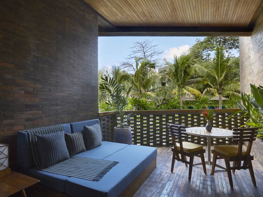 A modern outdoor seating area with a blue sofa and wooden chairs, a small table, and a view of lush greenery and palm trees.