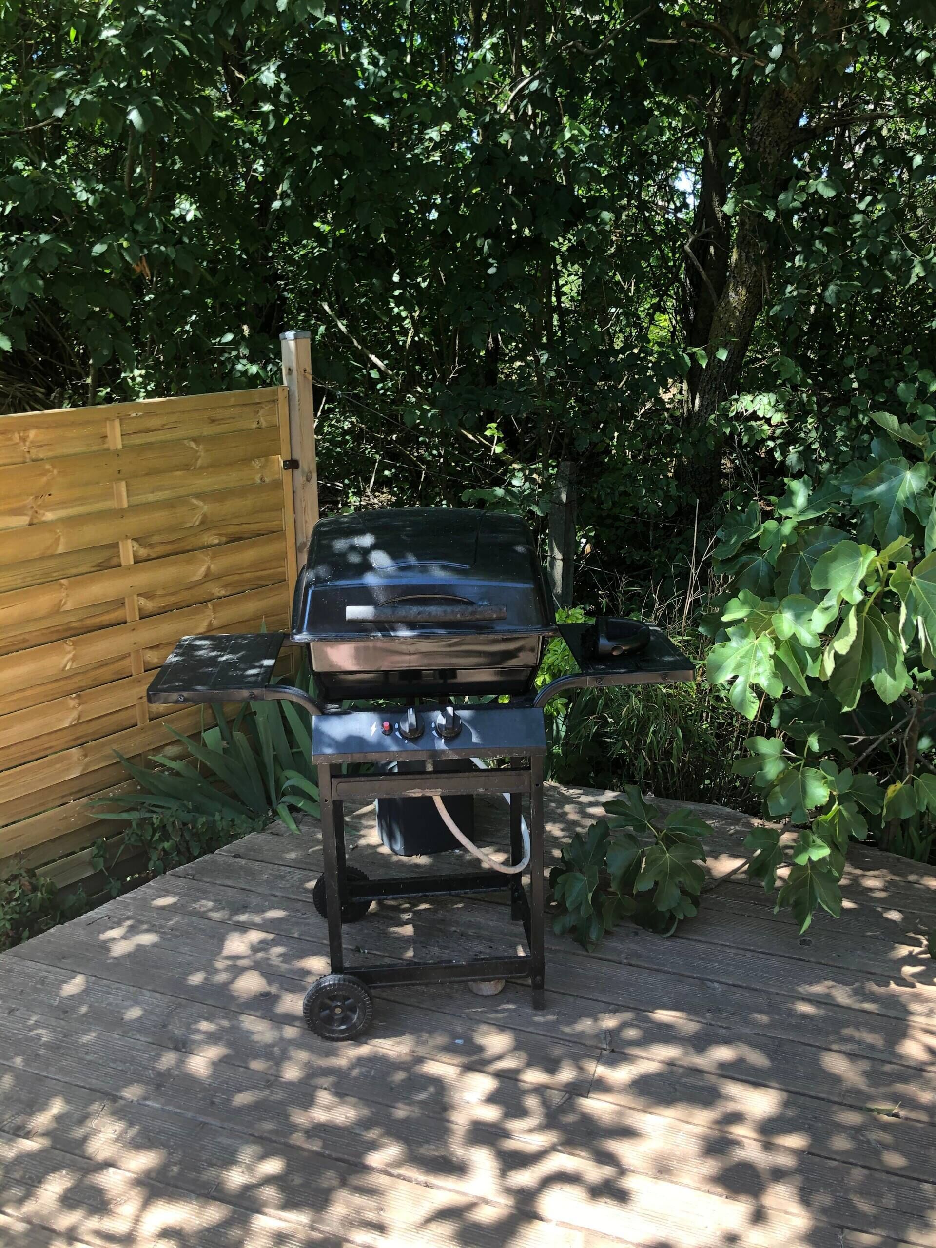 Ein tragbarer Grill auf einer Holzterrasse mit Holzzaun und Grünpflanzen im Hintergrund.