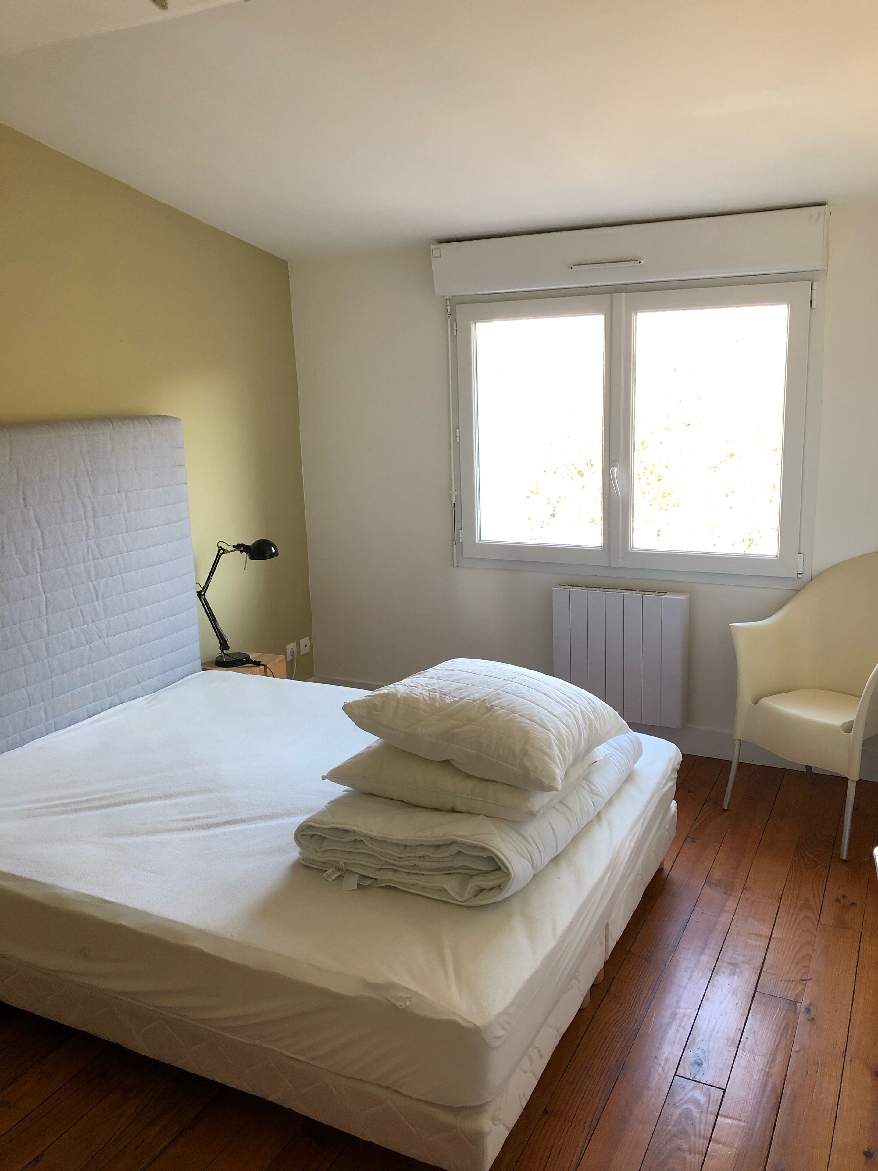 Ein ordentlich bezogenes Bett mit Kissen, einer Nachttischlampe, einem Stuhl und einem Fenster mit Jalousien.
