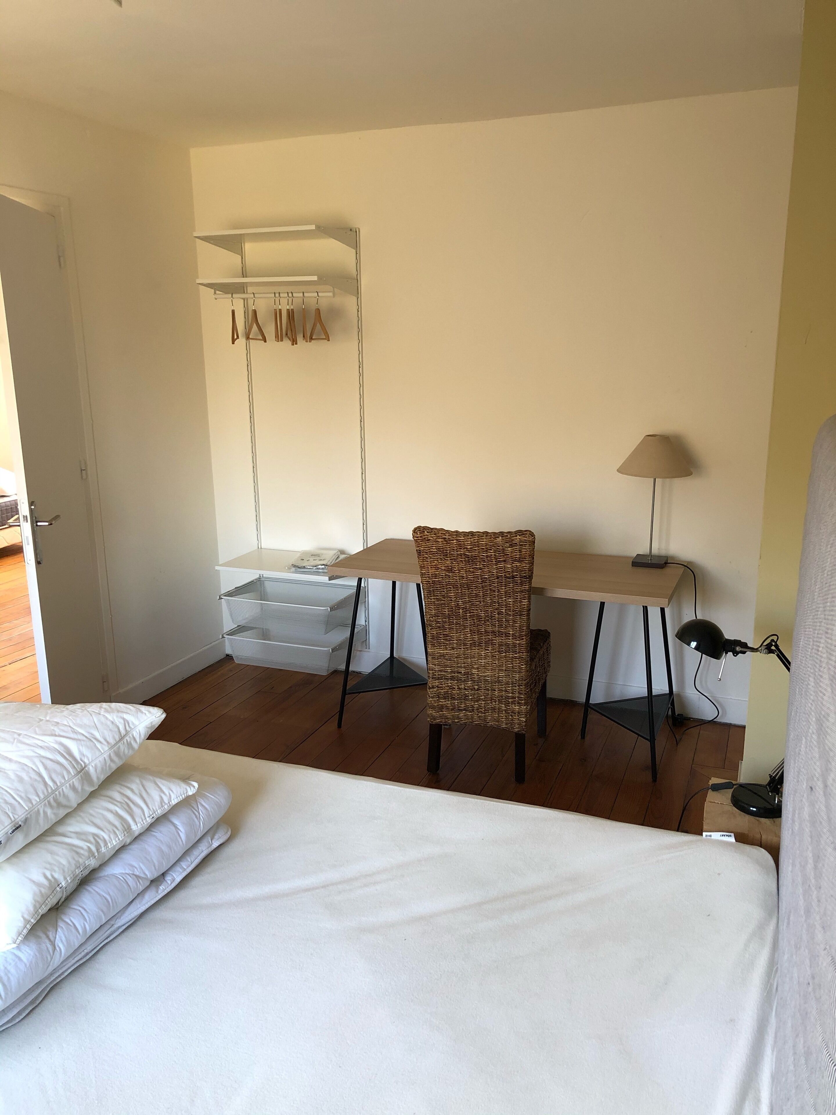 Une chambre d’hôtel équipée d’un lit, d’un bureau, d’une chaise, d’une lampe et d’une armoire où sont suspendus des vêtements.