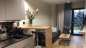 Ett modernt kök med en inbyggd brödrost, en köksö i trä och en kaffemaskin. Där finns en svart soffa med ett mönstrat kuddöverdrag, ett litet träbord och en vas med blommor. Ett stort fönster med gardiner syns, och det ger en fin utsikt över omgivningen.