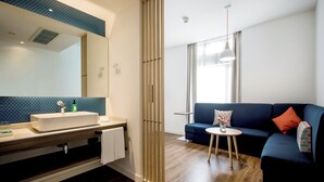 Une salle de bain moderne avec un lavabo, un miroir et un porte-serviettes. Le salon est équipé d’un canapé bleu et d’une petite table ronde avec une lampe.