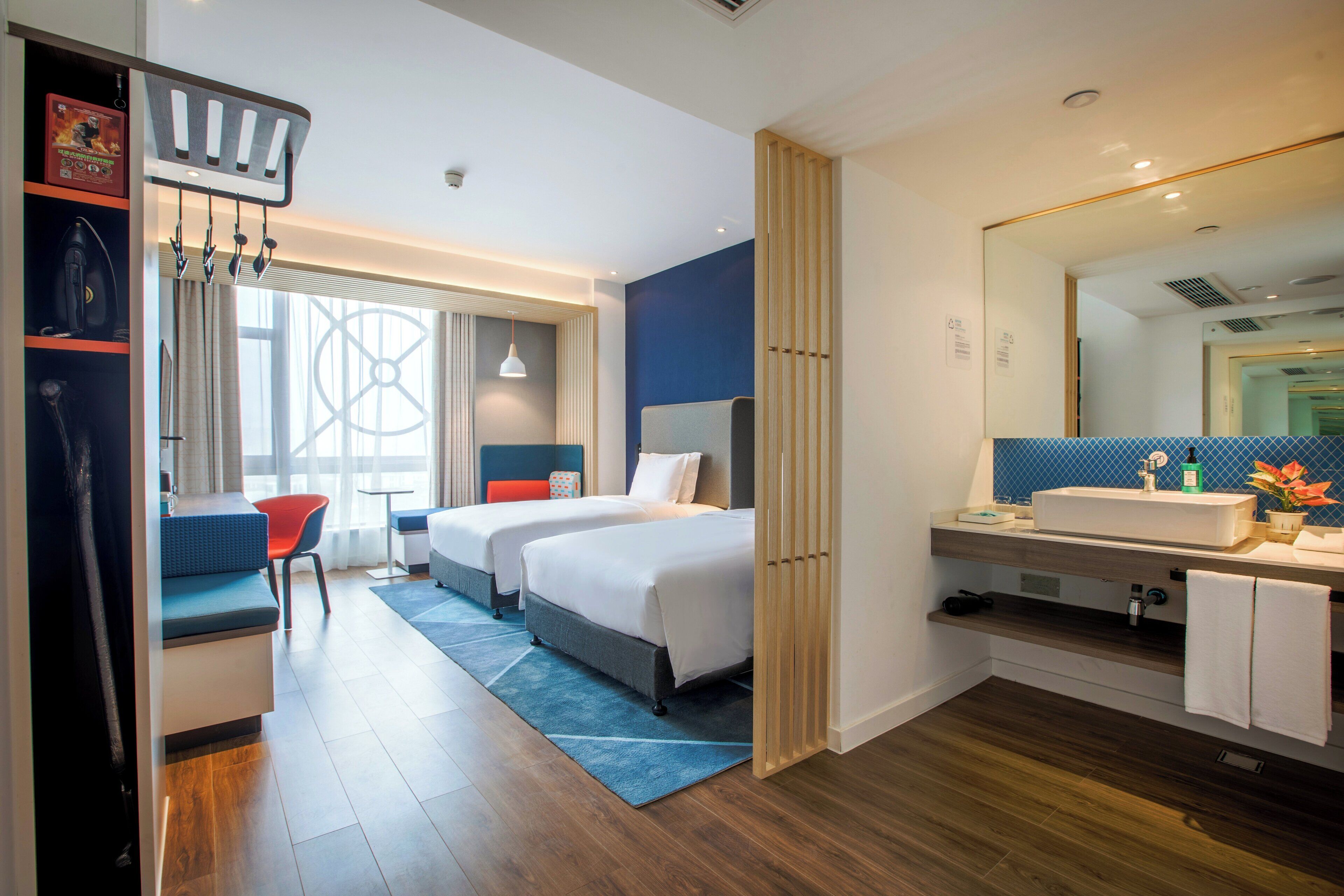 A hotel room with two beds, a desk, a chair, a bathroom with a mirror, and a window with curtains.