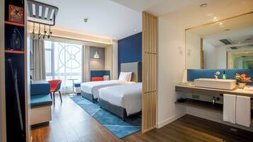A hotel room with two beds, a desk, a chair, a bathroom with a mirror, and a window with curtains.