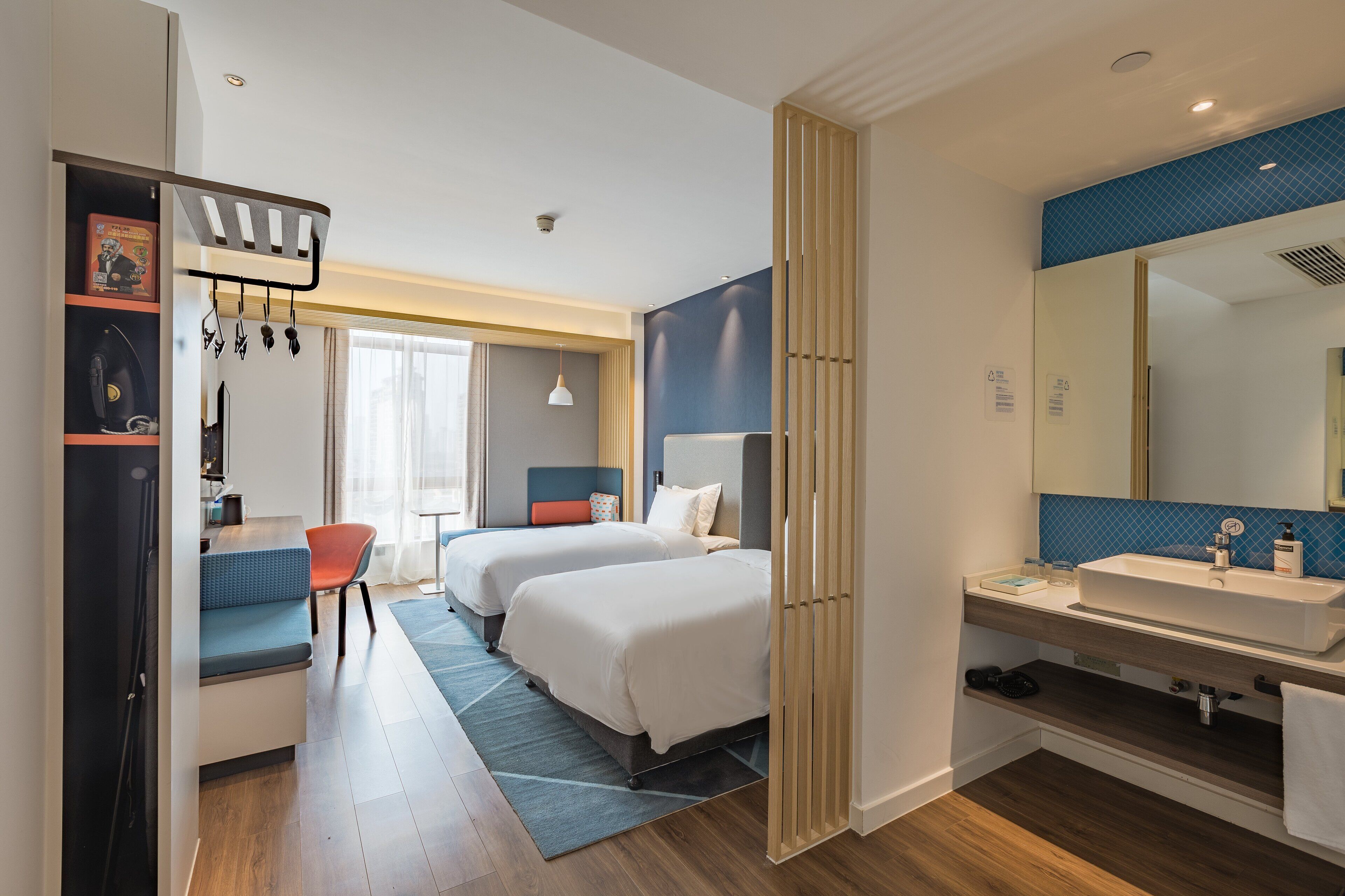 Camera d'albergo con due letti, una scrivania, una sedia, uno specchio grande e un bagno con lavandino e specchio.