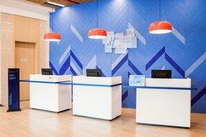 Un espace d’accueil avec trois comptoirs, chacun équipé d’un écran d’ordinateur et d’une enseigne indiquant « Holiday Inn Express ». Trois lampes rouges suspendues sont placées au-dessus des comptoirs, et le mur est orné d’un motif géométrique bleu.