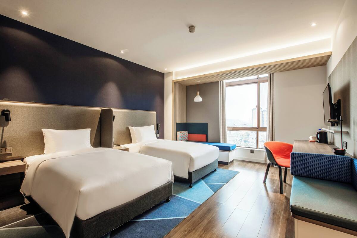 A hotel room with two beds, a desk, a chair, and a large window.