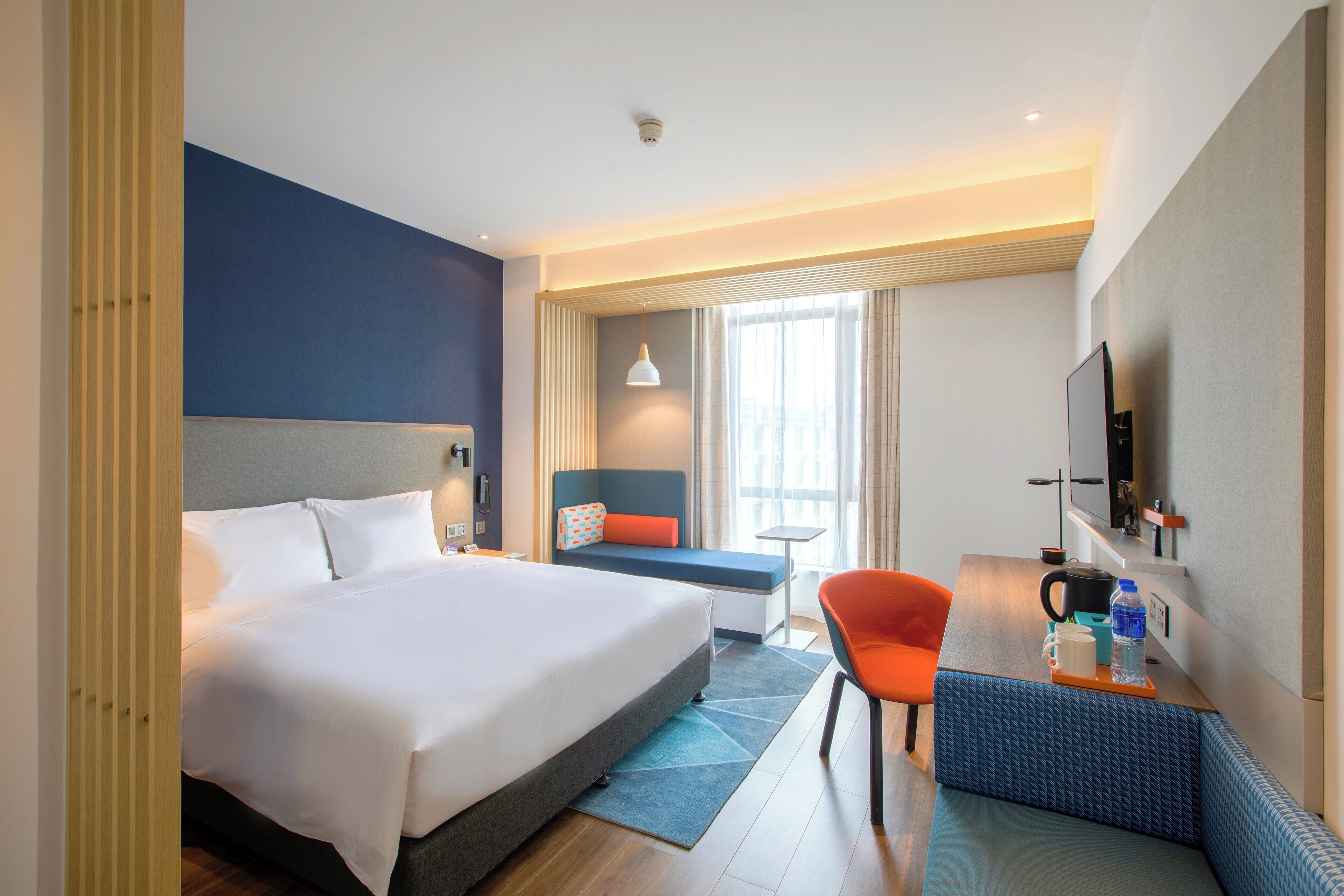 A modern hotel room with a large bed, a desk with a chair, a small table, and a television.
