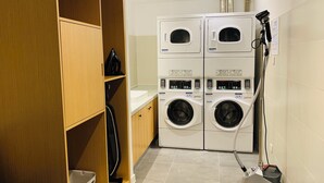A laundry room with stacked washing machines and dryers, a vacuum cleaner, and wooden shelving. - Holiday Inn Express Zhangjiagang Free Trade Zone by IHG (Suzhou)