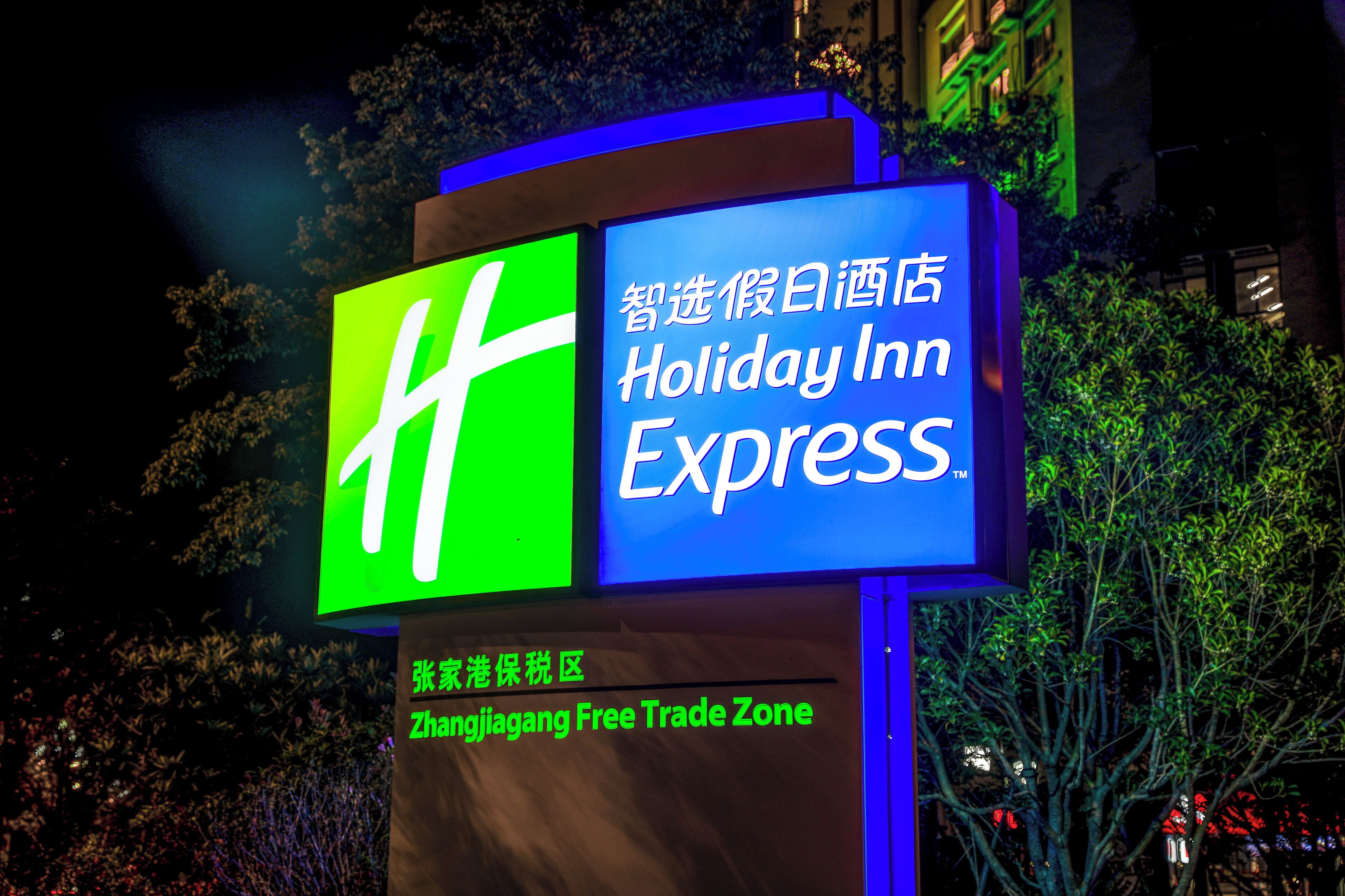 Ein Schild des Holiday Inn Express mit Grün- und Blautönen, gelegen in einem Geschäftsgebiet mit Bäumen und Gebäuden im Hintergrund.